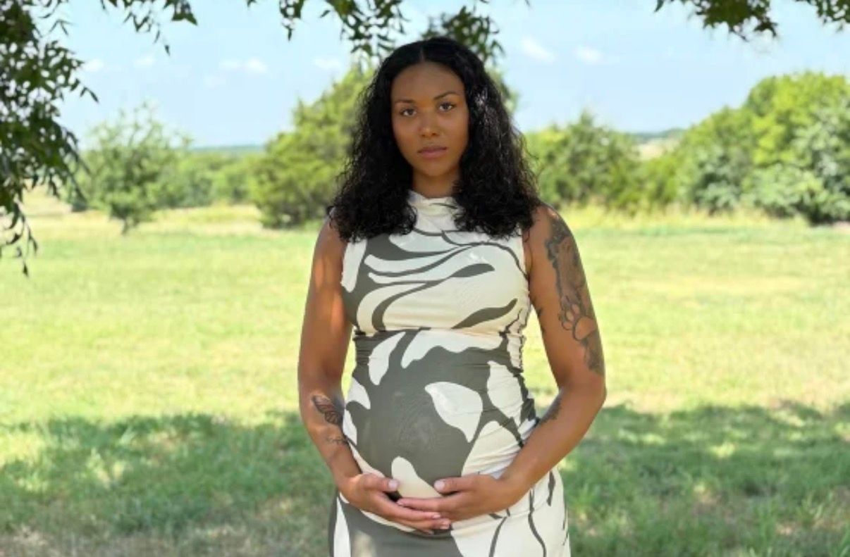 A pregnant woman standing in a grassy field, wearing a patterned dress and looking directly at the camera.