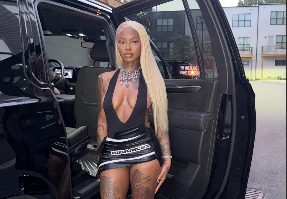 A woman with long blonde hair wearing a revealing black outfit stands in the open door of a black SUV.