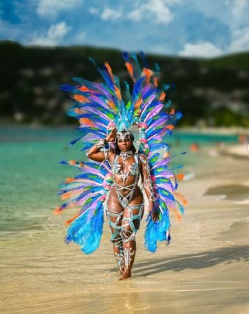 A person wearing a colorful feathered headdress and beaded costume walks on a sandy beach with a turquoise ocean in the background.