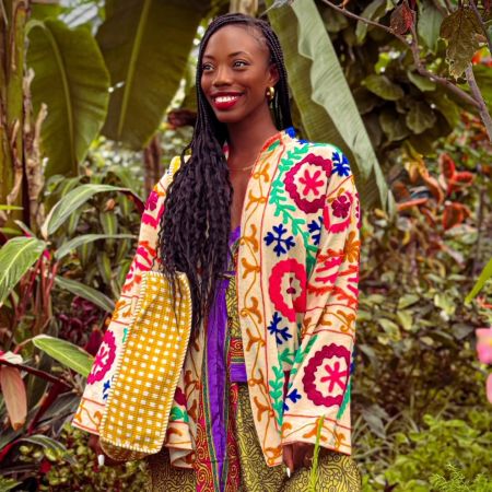 A smiling woman with long braided hair wearing a colorful, patterned jacket stands in a lush, tropical setting.