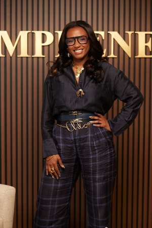 A smiling woman wearing glasses, a black shirt, and plaid pants stands in front of a backdrop with the word "EMPIRE" displayed.