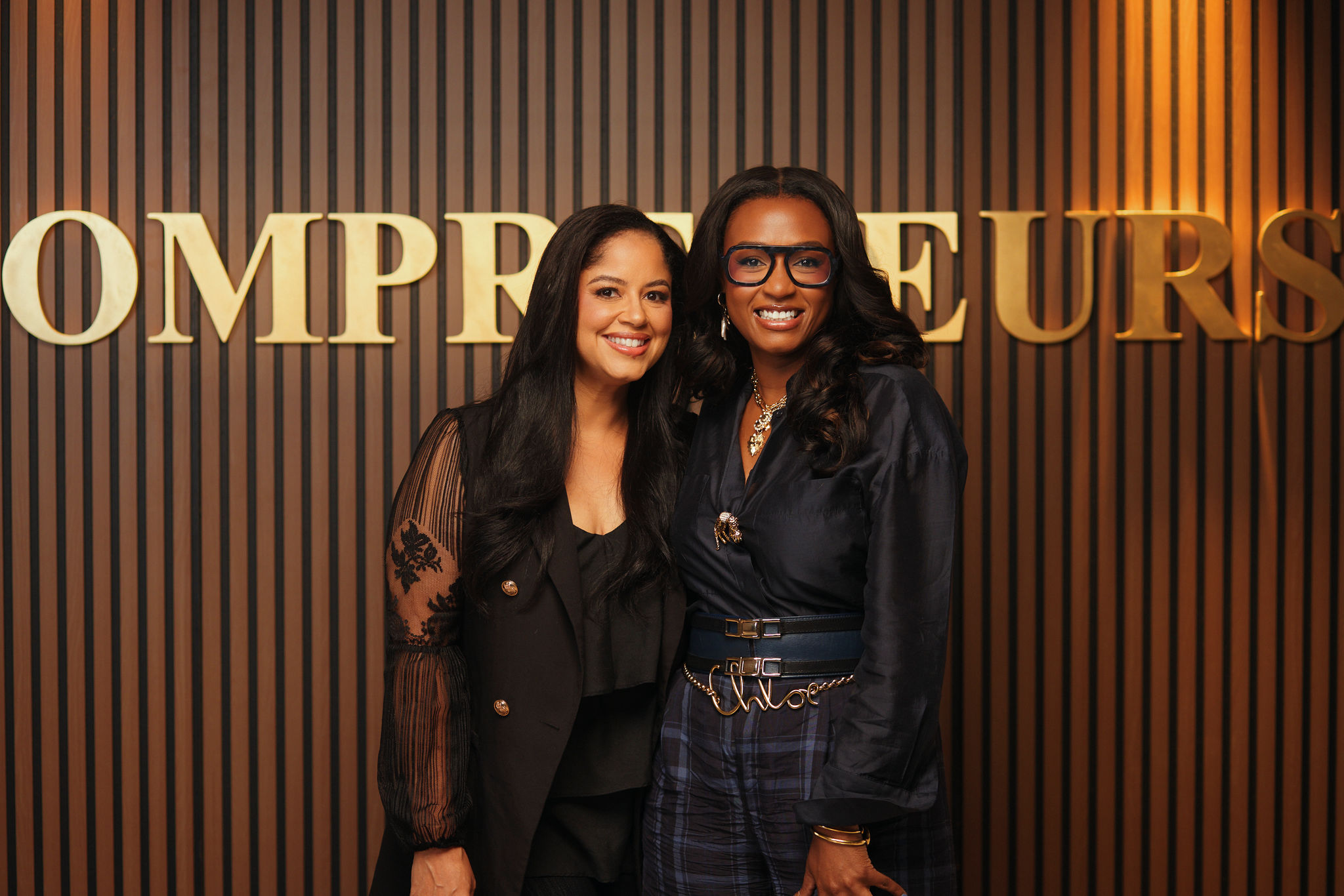 Two women posing in front of a large "Entrepreneurs" sign, one wearing a black outfit and the other wearing a black and white outfit.