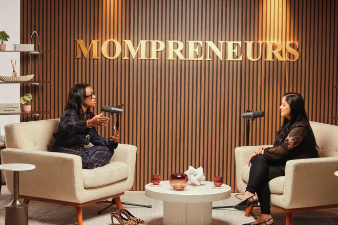 Two women sitting in chairs having a conversation in a room with "Mompreneurs" sign on the wall.