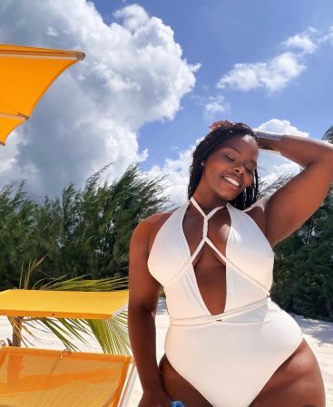 A smiling woman in a white swimsuit stands in front of a tropical setting with palm trees and a bright orange structure.