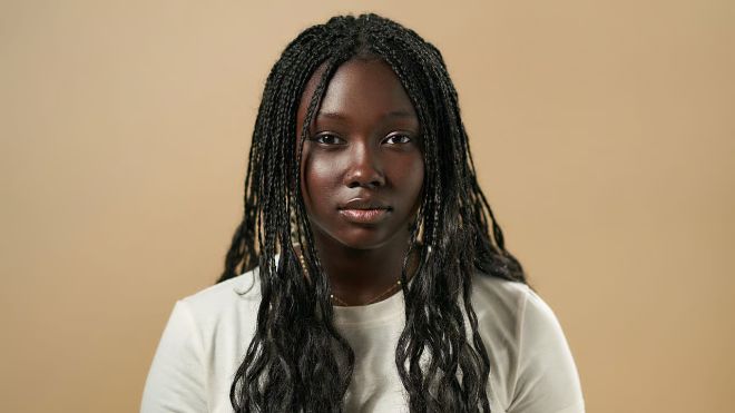 A young Black woman with long braided hair, wearing a white shirt, looking directly at the camera with a neutral expression.