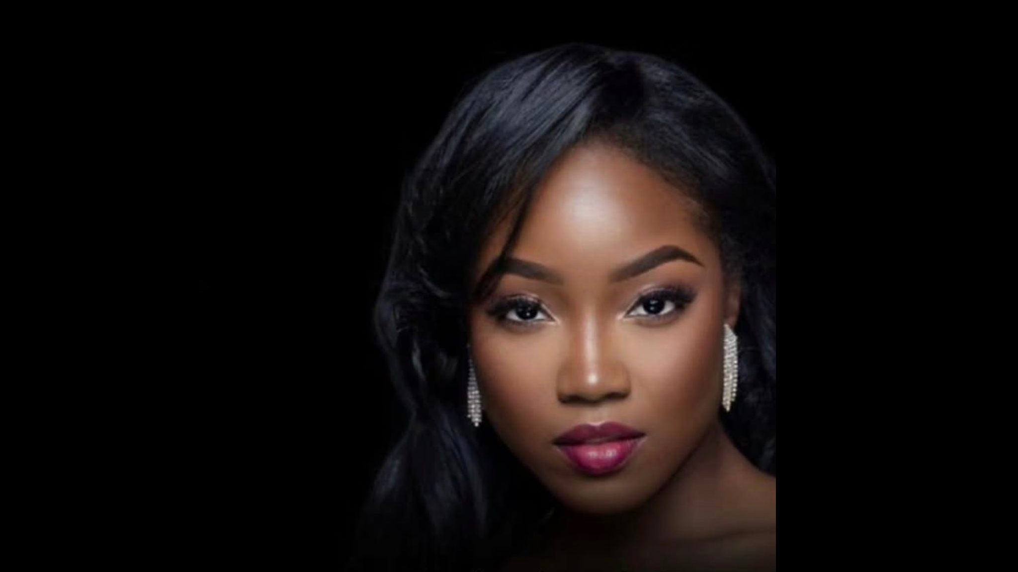 A close-up portrait of a woman with dark skin, long black hair, and red lipstick against a dark background.