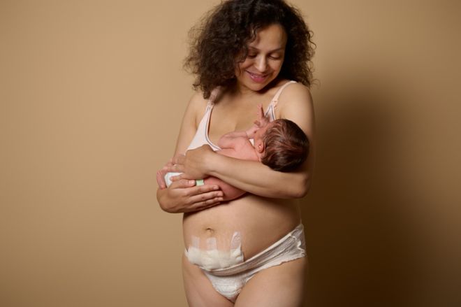 Authentic mom in underwear, smiles looking at her newborn baby she's holding, shows her body with postpartum scars flaws